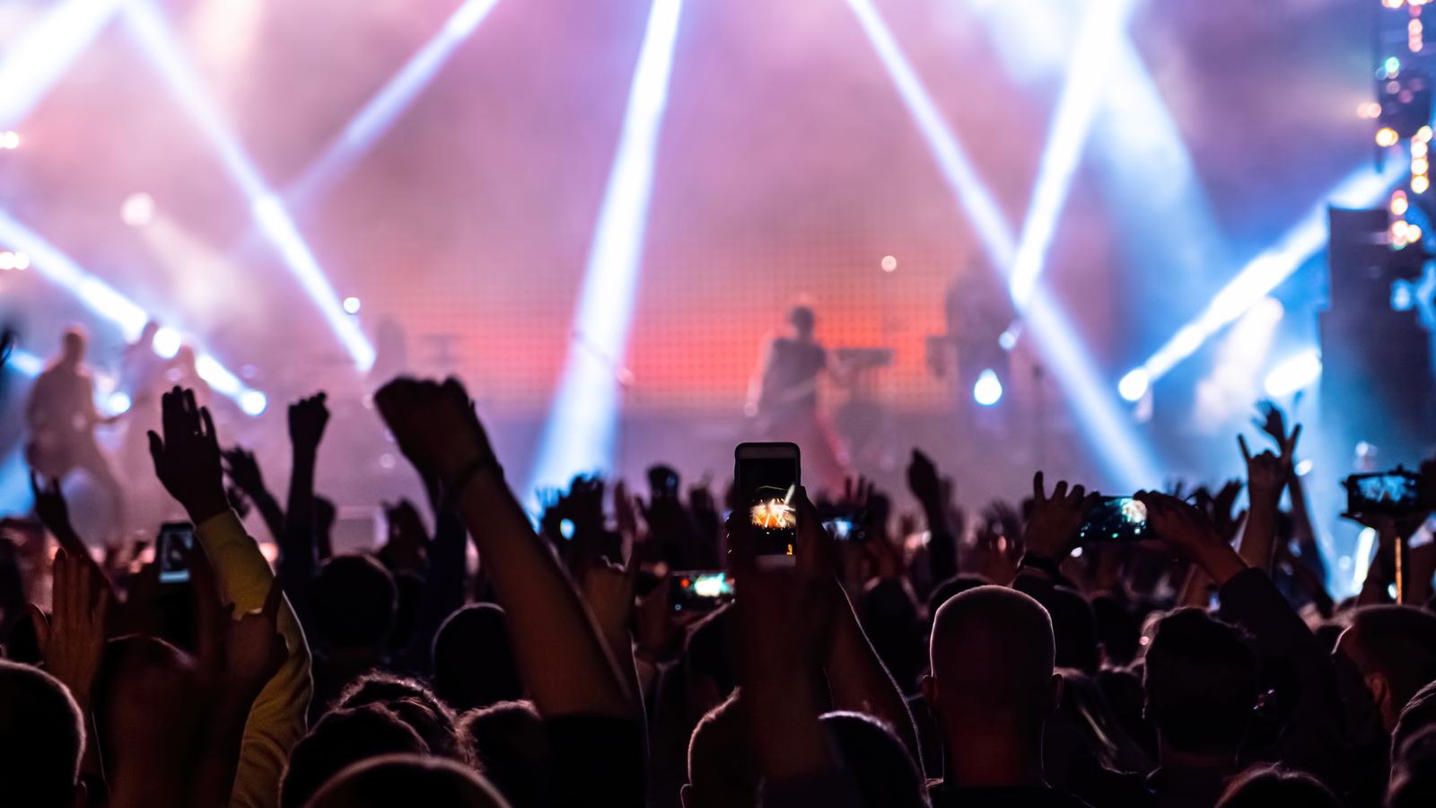 A packed concert crowd seen from behind, phones raised under red stage lights